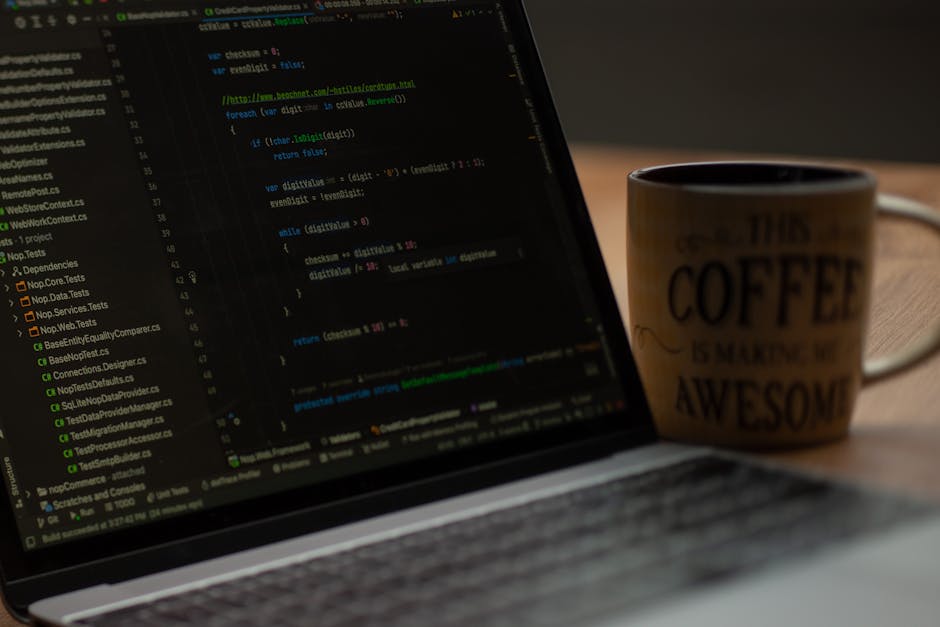 A close-up photograph showing a laptop with visible lines of colorful programming code on its screen, placed on a wooden desk. To the right of the laptop, there is a ceramic mug with a beige backgroun