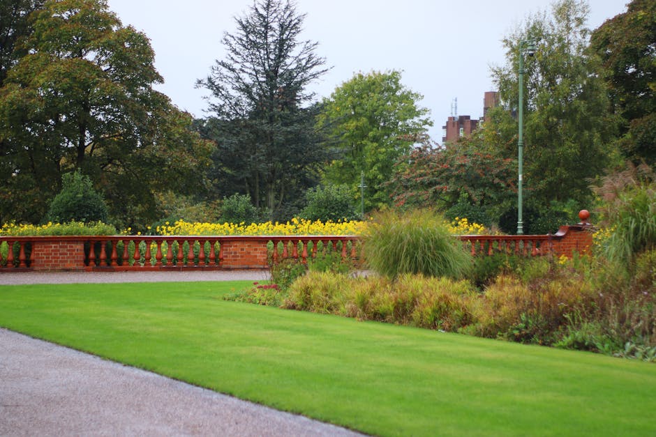 A well-maintained small front garden featuring a neatly trimmed lush green lawn with defined edges, bordered by a gravel pathway on the left. The garden includes a variety of ornamental plants and gra