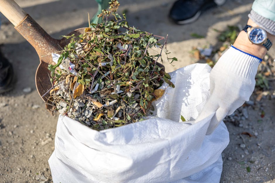 A person wearing a white glove and a wristwatch is using a small shovel to scoop up a pile of garden debris, including dead leaves, small branches, and soil, into a large white garden waste bag. The b