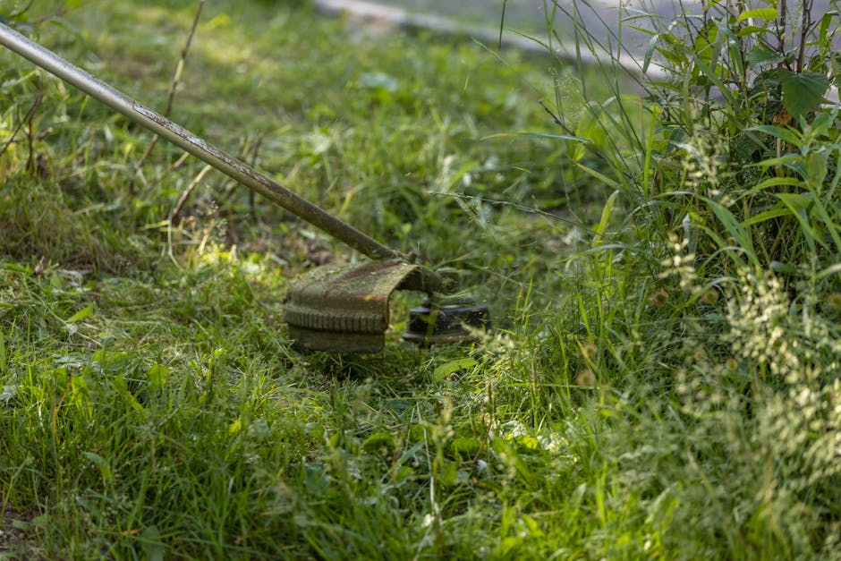 A person wearing a white glove and a wristwatch is using a small shovel to scoop up a pile of garden debris, including dead leaves, small branches, and soil, into a large white garden waste bag. The b
